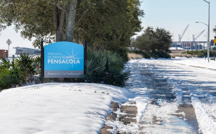 A blue sign stands on a snowy road, indicating "Downtown PENSACOLA America's 1st Settlement."