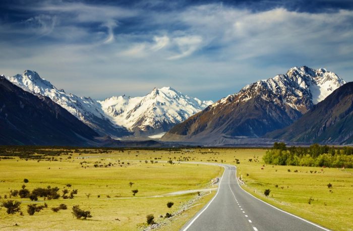 Scenic landscape with a winding road leading to snow-covered mountains under a partly cloudy blue sky.