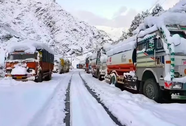 Line of colorful trucks stuck on a snow-covered mountain road surrounded by steep white peaks under a cloudy sky.