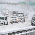 Cars and trucks driving slowly on a highway during a heavy snowstorm, with snow covering the road and vehicles.