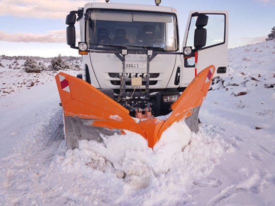 Snowplow truck clearing a road covered in deep snow in a rural or mountainous area.
