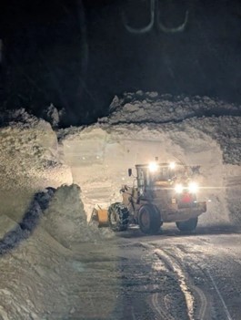 Heavy machinery clearing a large wall of compacted snow at night, illuminated by vehicle lights.