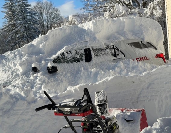 A vehicle almost entirely covered by a thick layer of snow, with snow removal equipment visible nearby.