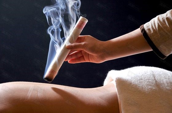 Close-up of a hand holding a smoking moxibustion stick near a person’s leg during a traditional heat therapy session.