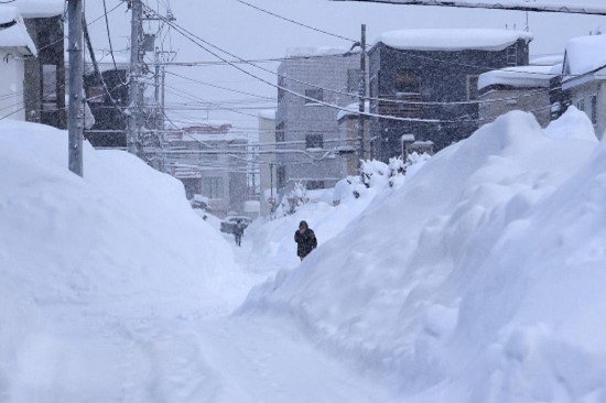 Urban street scene with extremely high snowbanks on both sides of a narrow cleared path, and a person walking through the snowy corridor.