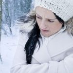 Woman wearing a winter coat and knit hat standing in a snowy forest, holding her arms as if feeling cold.