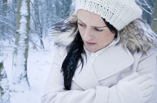 Woman wearing a winter coat and knit hat standing in a snowy forest, holding her arms as if feeling cold.