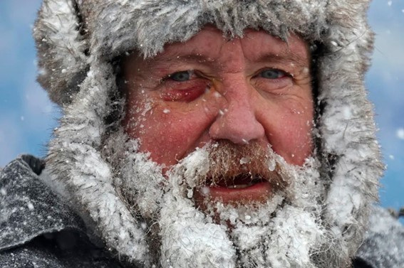 Close-up of a man outdoors in extreme cold, with frost covering his beard, mustache, and fur-lined hat.