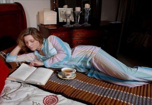 Woman lying on a bed reading a book while resting on a row of cylindrical heat packs, with a cup of tea placed beside her.