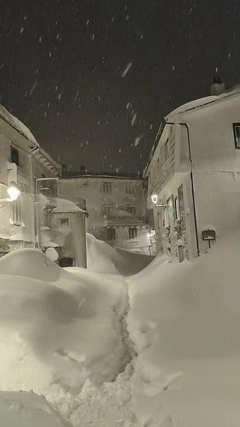Narrow street heavily covered in deep snow between buildings at night.